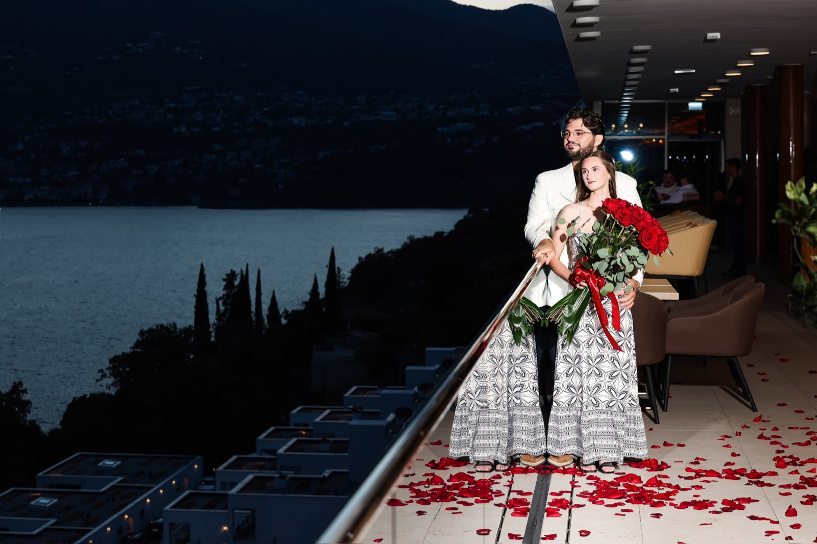 Tatum and Zachary standing together with red roses overlooking the water at night