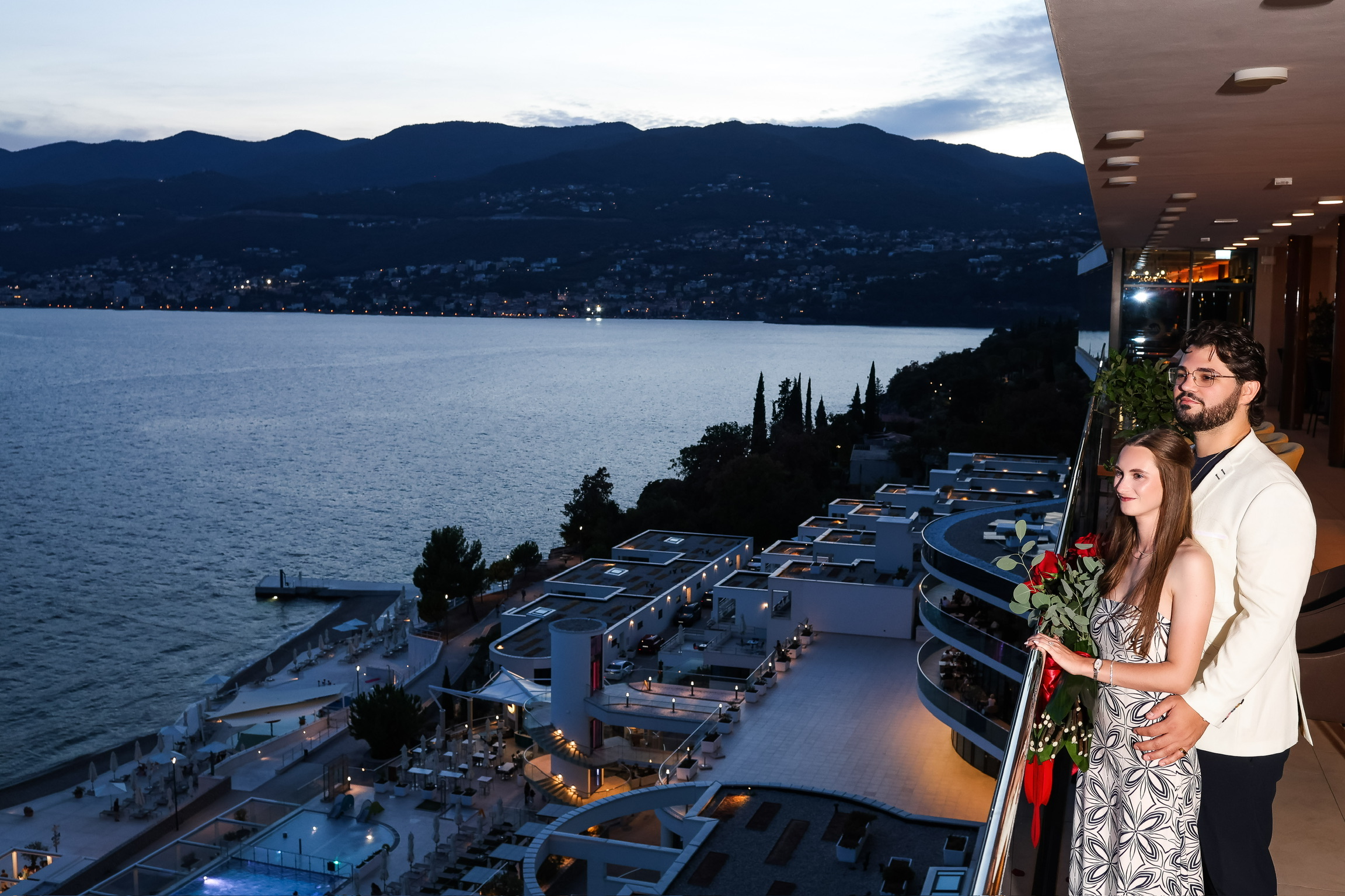 Tatum and Zachary embracing on a balcony overlooking the water at dusk